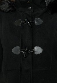 Črn volnen plašč s ovratnikom iz umetnega krzna, opremljen s preklopnimi zapirali in gladkimi usnjenimi detajli. Tekstura blaga pripomore k toplini.