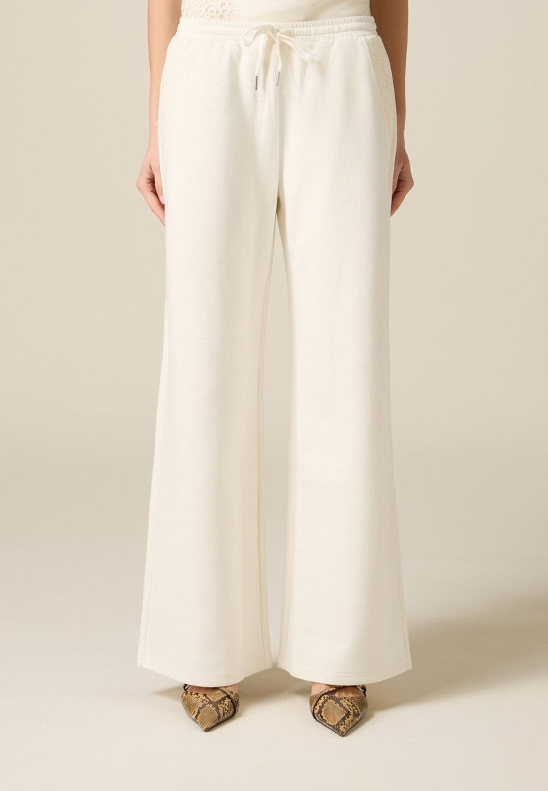 Kobieta w białych spodniach o szerokich nogawkach z wiązaniem w pasie, nosząca buty wężową wzorem i zakończone ostrym noskiem, stojąca na neutralnym tle.