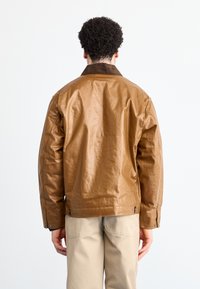 Giacca sintetica marrone con finitura liscia, collo in corduroy e chiusure a scatto sui polsini. Indossata sopra pantaloni beige, mostrata da dietro.