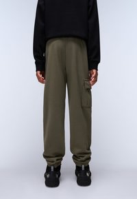 Olivgrüne Sweatpants mit lockerer Passform, elastischem Bund und seitlicher Cargo-Tasche. Ein Paar schwarze Sneakers mit strukturierten Sohlen.