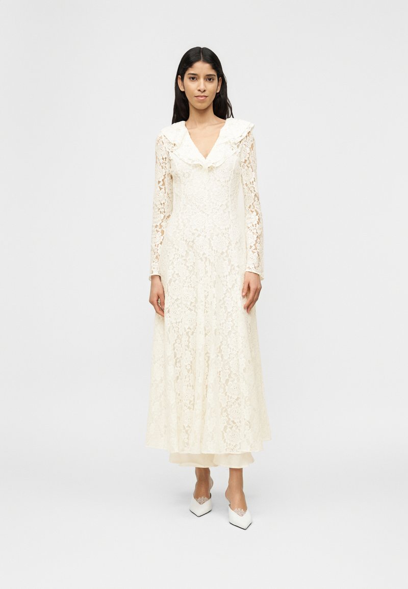 Γυναίκα που φοράει μακρυμάνικο, ολ όσωμο λευκό φόρεμα από δαντέλα με V-λαιμόκοψη με βολάν και λευκά παπούτσια με μυτερή μύτη, στέκεται μπροστά σε ένα απλό φόντο.