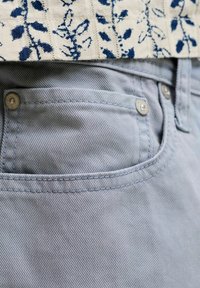 Σκούρο γαλάζιο τζιν τζιν με μπροστινή τσέπη με ορατή ραφή και μεταλλικά πριτσίνια, συνδυασμένο με λευκό ύφασμα με μπλε ανθισμένα σχέδια.