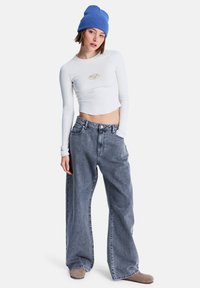 Λευκό ριμπ μακρυμάνικο crop top με χρυσό λογότυπο, σε συνδυασμό με φαρδιά παντελόνια από γκρι τζιν και μπλε πλεκτό σκουφάκι. Ταμπά παπούτσια.