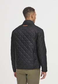 Svart, quiltet jakke med stående krage, strukturert overflate og et brunt lærdetalj på baksiden av skuldrene. Klassisk snitt og design.