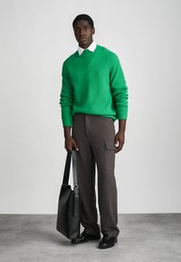 Πράσινος ριγέ πουλόβερ, λευκό πουκάμισο με γιακά, καφέ παντελόνι με τσέπη στο πλάι, μαύρη τσάντα. Το μοντέλο στέκεται σε γκρι πάτωμα εναντίον ενός ανοιχτού φόντου.