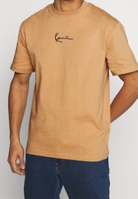 Camiseta de manga corta y cuello redondo en algodón marrón claro con un logo en cursiva negro en el pecho. Textura suave y corte relajado.