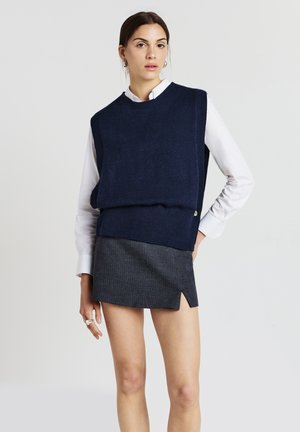 Marineblauer Strickpullover über einem weißen Kragenhemd, kombiniert mit einem grauen Miniröckchen mit einem Seitenschlitz und vertikalen Nadelstreifen.