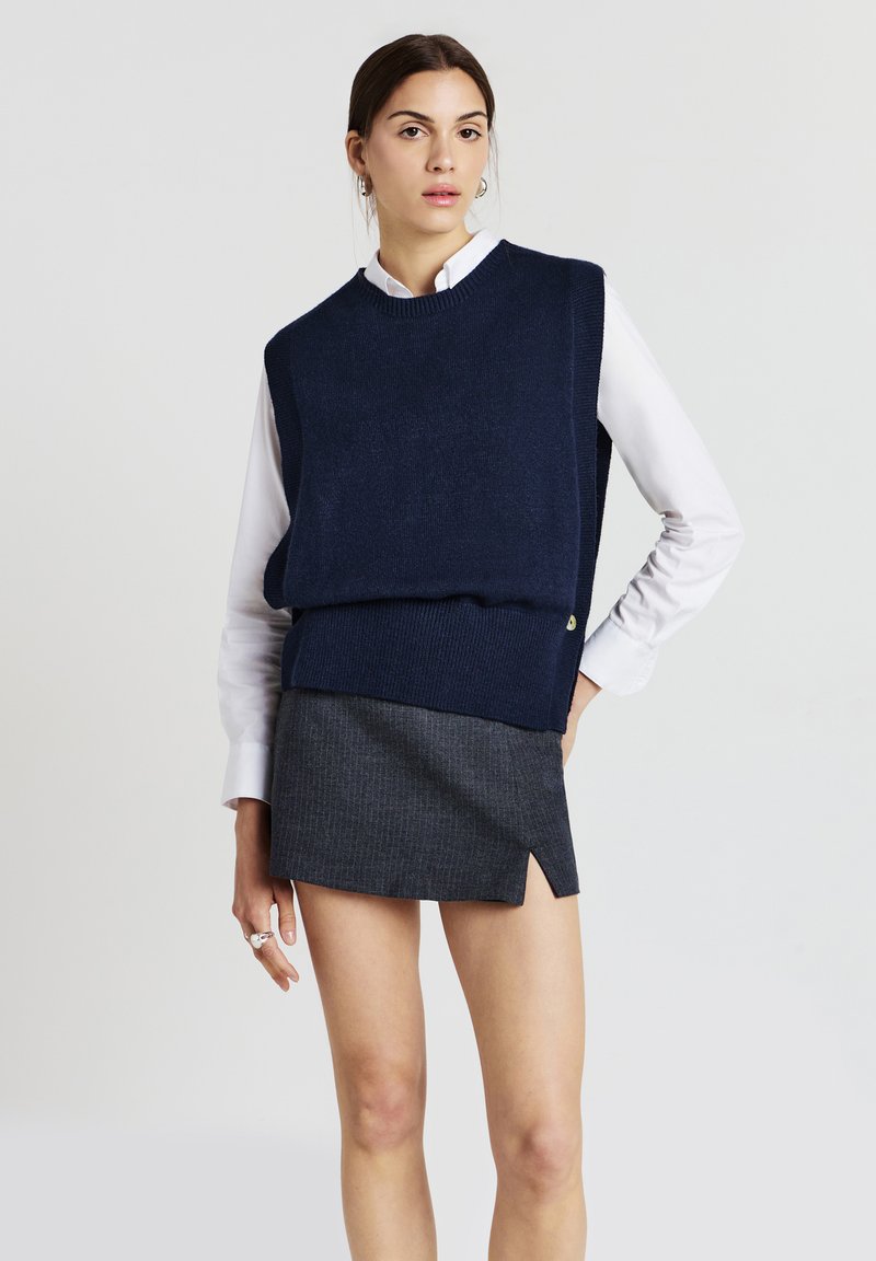 Gilet en tricot bleu marine porté sur une chemise blanche à col, associé à une mini-jupe grise dotée d'une fente latérale et de rayures verticales.