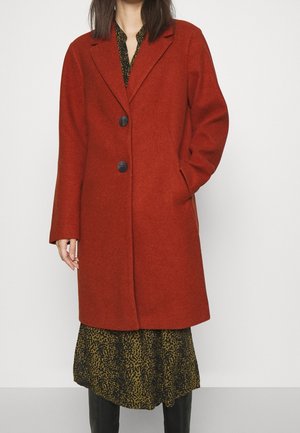 Femme portant un manteau en laine couleur rouille avec des boutons noirs sur une robe à motifs verts et noirs et des bottes noires montant jusqu'aux genoux.