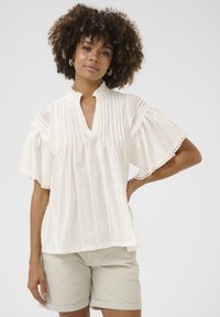 Blusa bianca con maniche corte a campana; pieghe e motivi testurizzati; colletto arruffato; indossata con pantaloni corti chiari.