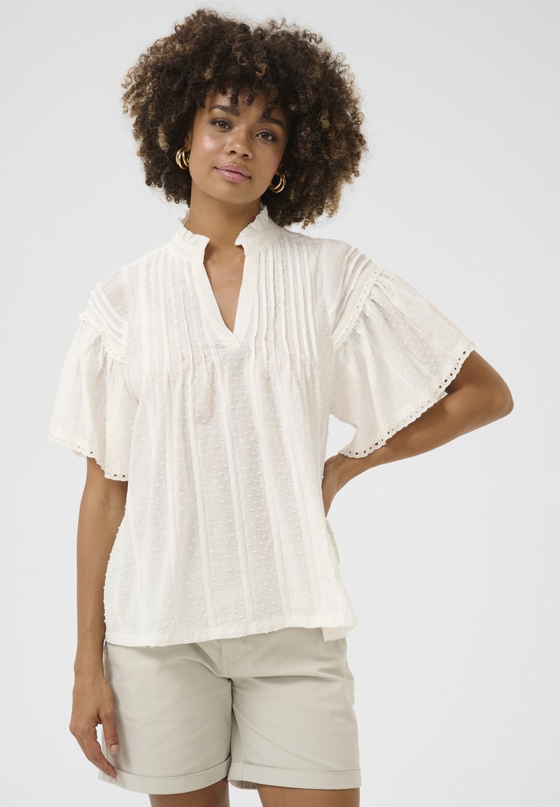 Blusa bianca con maniche corte a campana; pieghe e motivi testurizzati; colletto arruffato; indossata con pantaloni corti chiari.