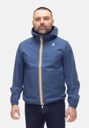 Uomo che indossa una giacca con cappuccio blu con una cerniera a strisce gialle e bianche, in piedi davanti a uno sfondo bianco uniforme.