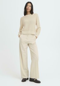 Mujer vestida con un suéter de punto beige, pantalones de pierna ancha crema y mocasines negros, de pie con las manos en los bolsillos frente a un fondo claro.