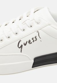 Sneaker bianco con tomaia in ecopelle testurizzata, lacci bianchi piatti, suola in gomma nera e scritta a mano "Guess?" di lato.