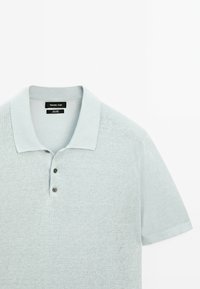 Světle modré polo triko s krátkým rukávem z jemného pleteného materiálu, s plochým límečkem, zapínáním na tři knoflíky a jemnou texturou.