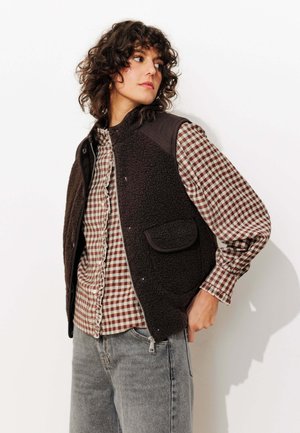 Gilet en polaire marron avec fermeture éclair et poche latérale, superposé à une chemise à carreaux marron et blanc à boutons. Un jeans gris clair complète le look.