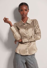 Beige satijnen shirt met een puntige kraag, lange mouwen en knoopsluiting aan de voorkant. Gladde textuur en slim fit, gecombineerd met grijze gestreepte broek.