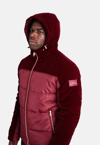 Giacca cappuccio bordeaux con design trapuntato, texture a contrasto, accessori in oro e un'etichetta sulla manica sinistra.