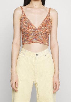 Frau trägt ein mehrfarbiges gestricktes Crop-Top mit dünnen Trägern und hoch taillierte, blassgelbe Cordhosen, steht vor einem schlichten Hintergrund.