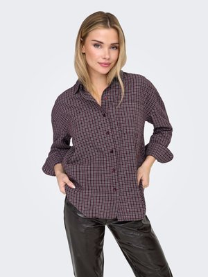 Chemise à carreaux boutonnée de couleur rouge foncé et bleu marine, manches longues retroussées. Associée à un pantalon en similicuir noir brillant.