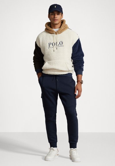 Mikina z fleece v krémovej, námorníckej a hnedej farbe s šnúrkami a logom. Spárované s námorníckymi teplákmi a bielymi teniskami, ktoré majú bočné vrecká.