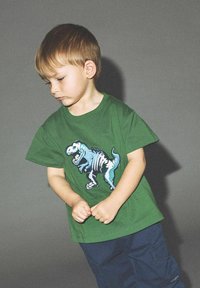 Roheline T-särk sinise ja valge dinosauruse graafikaga. Lühikesed varrukad, ümar kaelus. Laps kannab tumedaid siniseid lühikesi pükse, seistes käed koos.