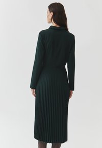 Abito plissettato verde scuro con maniche lunghe e colletto. Il tessuto è liscio, mettendo in evidenza pieghe verticali dalla vita all'orlo.