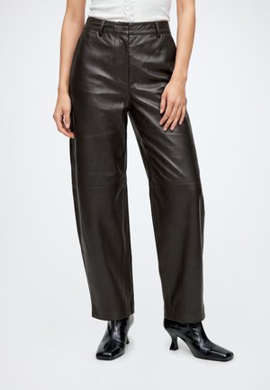 Femme portant un pantalon en cuir noir taille haute à jambes larges, associé à des bottines noires à talons, sur fond blanc.