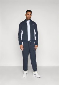 Homem de pé a usar um fato de treino azul-marinho e branco, com um casaco com fecho e ténis brancos, contra um fundo claro e liso.