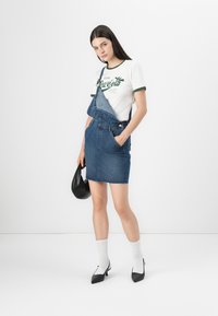 Denimist üldkleid, millel on kitsas seelik ja kulunud äär. Koos valge graafilise T-särgiga roheliste aktsentidega ja musta nahkrahakotiga.