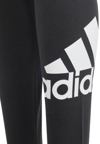 Pantalones deportivos negros de tela suave con un gran logo blanco de Adidas en el muslo izquierdo. El diseño incluye una forma ajustada y estilizada.