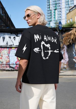 Person mit hellblonden Haaren und Sonnenbrille trägt ein schwarzes Shirt mit weißem "Mi Amor"-Text und einem von einem Pfeil durchbohrten Herz und steht auf einer städtischen Straße mit Graffiti.