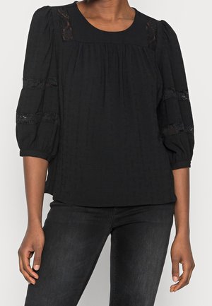 Femme portant un chemisier noir texturé avec des inserts en dentelle sur les épaules et les manches, associé à un jean noir délavé foncé.