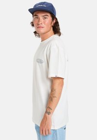 Hvid t-shirt med korteærmer og detalje med lyseblå logo, bæres med lyseblå jeans. En marineblå kasket supplerer outfittet.