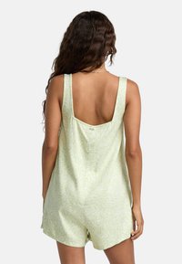 Grön mönstrad jumpsuit med djup rygg, breda axelband och lös passform. Materialet verkar vara slätt med en subtil blommig design.
