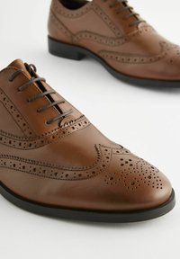 Chaussures oxford en cuir marron avec des détails de brogue perforés, bout arrondi, semelle en caoutchouc noire et lacets foncés. Texture lisse, design classique.