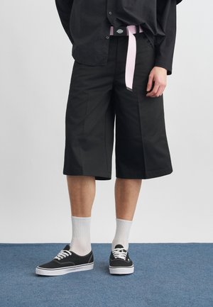 Persoon die zwarte, wijd uitlopende, knielange shorts draagt, een zwart shirt, een roze riem, witte sokken en zwarte lage sneakers, staand op een blauw tapijt.