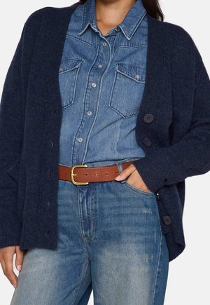 Kvinde iført en mørkeblå cardigan over en knappet denimskjorte, der er stoppet ned i lyseblå jeans med et brunt bælte.