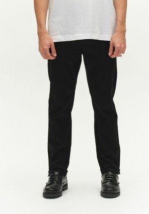 Persoon draagt zwarte broek met rechte pijpen, zwarte veterschoenen en een wit overhemd, staand tegen een effen lichte achtergrond.