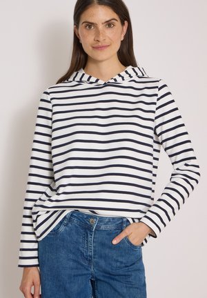 Kvinde med langt mørkt hår iført en hvid og marineblå stribet langærmet hættetrøje og blå jeans med den ene hånd i lommen.
