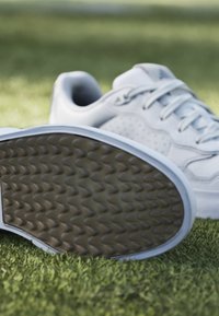 Vita sportskor med texturerade gummisulor och perforerade detaljer. Ovandelen har slät läder. Fångad på grön gräs.