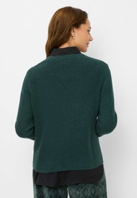 Donkergroene geribbelde gebreide trui met een ronde hals en lange mouwen, gecombineerd met een zwart overhemd met kraag, gezien vanaf de achterkant.