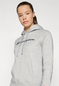 Pilka sportinė striukė su priekiu kišenėje, raišteliais ir ryškiai matoma "Champion" logo tamsiai mėlynos spalvos. Matoma minkšta audinio tekstūra.