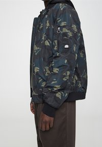 Bomberka w camo w odcieniach granatu i zieleni, wykonana z błyszczącej tkaniny, z zamkiem błyskawicznym, ściągaczami przy mankietach oraz kieszenią na ramieniu z detalem logo.