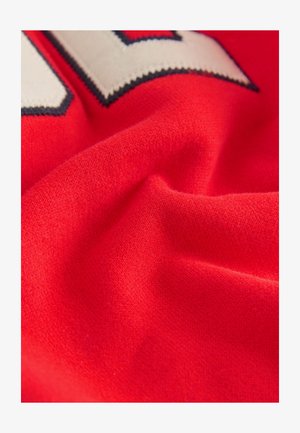 Tissu rouge à texture douce, présentant un motif blanc et bleu marine. Le matériau semble extensible et lisse, avec de subtiles variations dans le tissage.