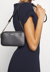 Borsa a tracolla nera DKNY con finitura texture, chiusura con zip e tracolla con catena, indossata sulla spalla su un vestito blu navy.