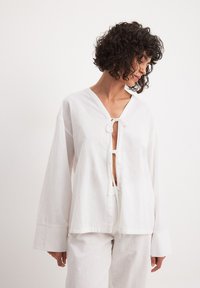 Blusa in lino bianco con scollatura a V, chiusura con lacci e maniche ampie. Tessuto morbido, materiale leggero e vestibilità comoda.