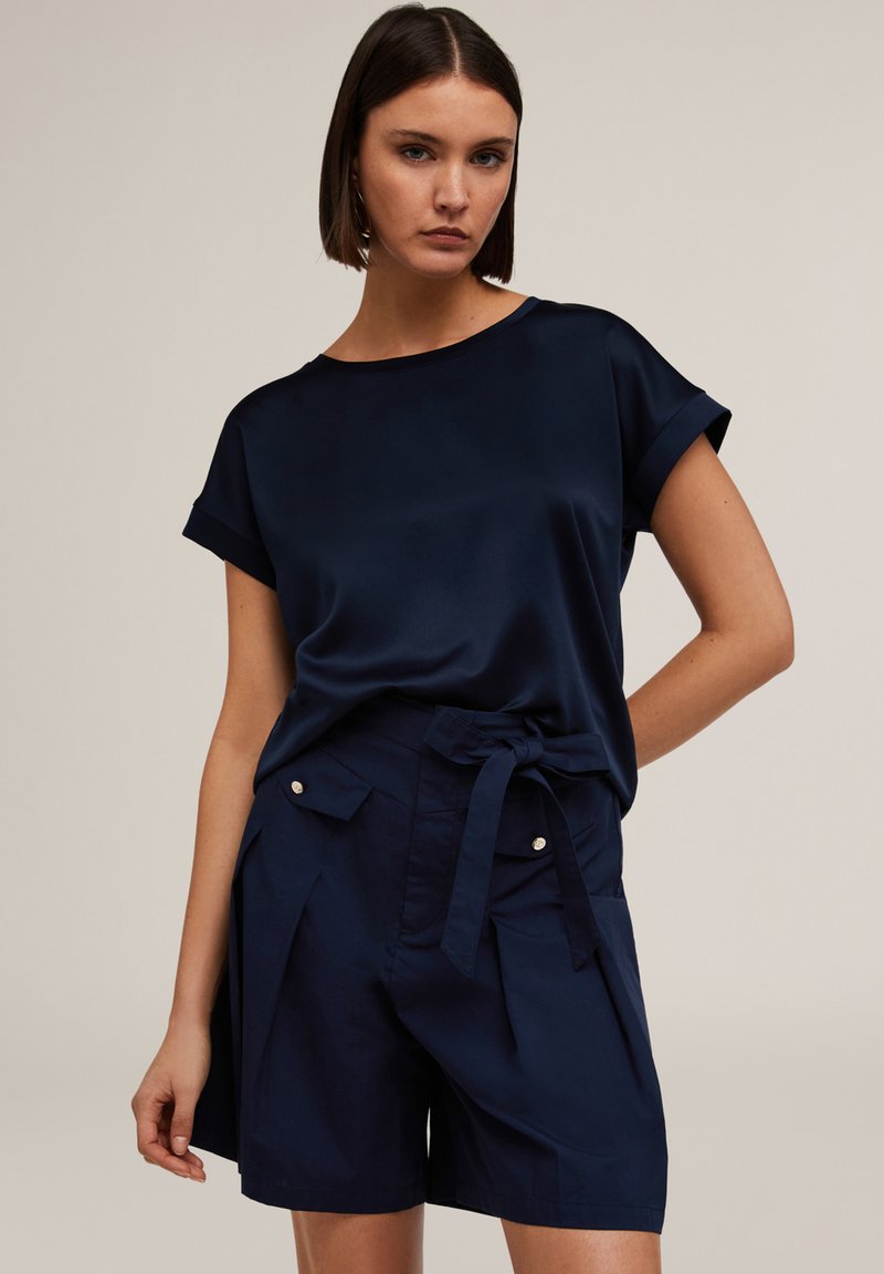 Top din satin de culoare albastru marin cu mâneci scurte și răsucite, asortat cu pantaloni scurți în nuanță de albastru marin, cu talie legată și buzunare frontale cu accente argintii.