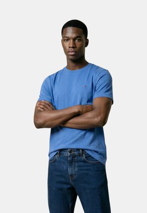 Mladý muž se založenýma rukama, oblečený v modrou košili s krátkým rukávem a tmavě modré džíny, stojící před jednobarevným světlým pozadím.