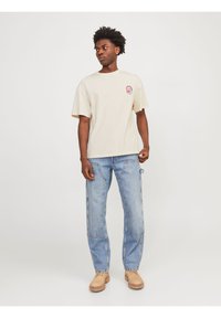 Beige bomulds T-shirt med et lille grafik på brystet, parret med løse blå denim jeans og lysebrune sko. Simpelt og afslappet design.